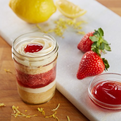 A layered dessert in a jar with lemon, strawberry, and cream, garnished with fresh strawberries and lemon slices.