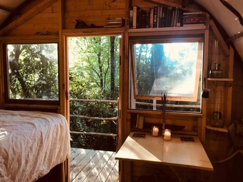 Cozy interior of a wooden cabin with large windows, a bed, and a small table with candles, overlooking a forest.