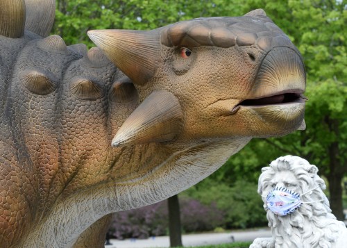 A large dinosaur statue with detailed textures stands next to a lion statue wearing a face mask in a park setting.