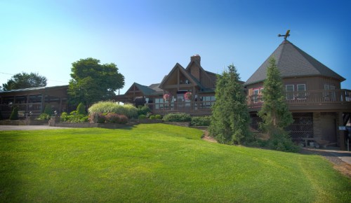 A large wooden lodge with a sloped roof, surrounded by green grass and trees under a clear blue sky.