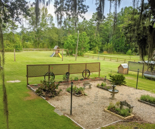 A serene backyard with a garden, playset, and lush green lawn surrounded by trees and a wooden fence.