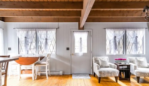 Bright, cozy interior with wooden beams, a round table, white chairs, and floral accents near large windows.