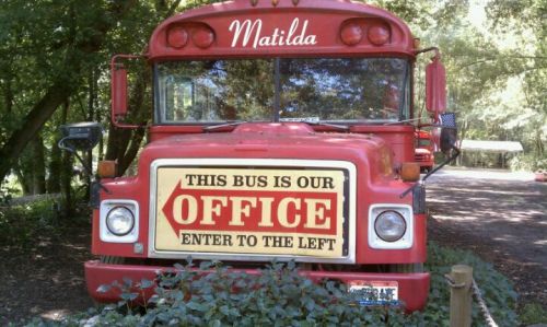 A red bus named "Matilda" with a sign reading "This bus is our office, enter to the left," surrounded by greenery.