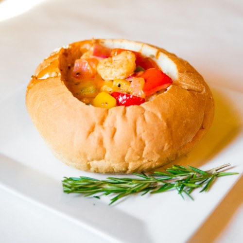 A bread bowl filled with colorful shrimp and vegetable stew, garnished with a sprig of rosemary on a white plate.