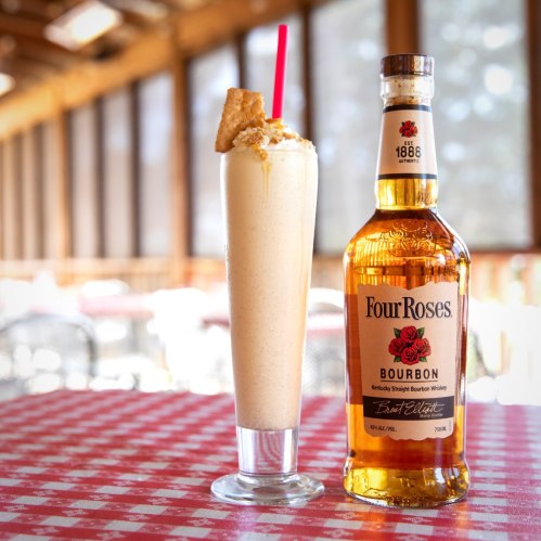 A tall glass of creamy bourbon cocktail with a red straw, next to a bottle of Four Roses Bourbon on a checkered tablecloth.