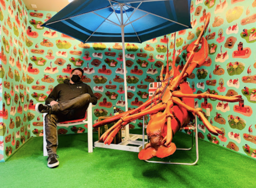 A person in a mask sits in a chair under an umbrella next to a large lobster sculpture in a colorful, patterned room.