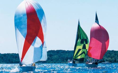 Three sailboats with colorful spinnakers sailing on a bright blue lake under a clear sky.