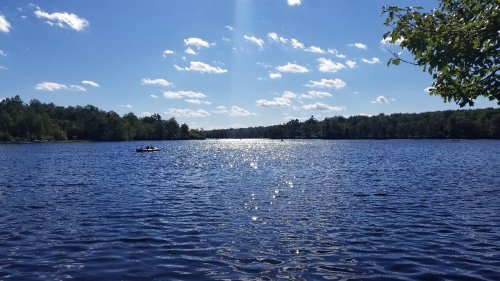 A serene lake under a bright blue sky, with fluffy clouds and sunlight reflecting on the water's surface.