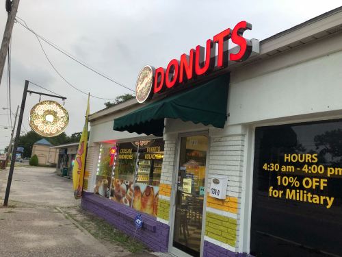 A donut shop with a bright red sign, featuring a green awning and promotional hours displayed on the window.