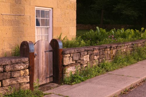 A small wooden gate in a stone wall beside a sidewalk, with greenery and flowers in the surrounding area.