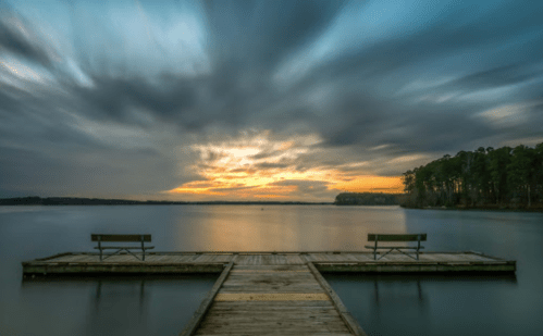 A serene lake view at sunset, featuring a wooden dock with two benches and dramatic, swirling clouds above.