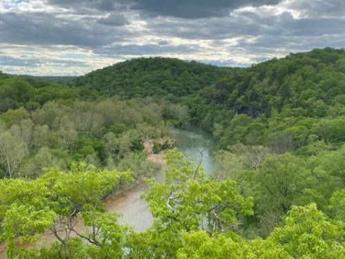 A scenic view of a winding river surrounded by lush green hills and trees under a cloudy sky.