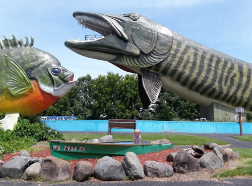 A child sits in a small boat surrounded by giant fish sculptures in a colorful outdoor setting.
