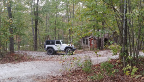 A white Jeep parked on a gravel path near a rustic cabin surrounded by trees in a wooded area.