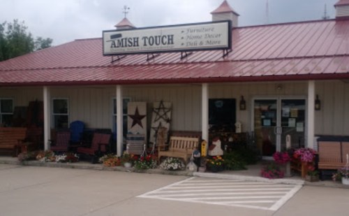 Exterior of a store named "Amish Touch," featuring furniture and home decor, with flower beds and a red roof.