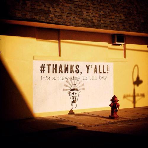 A mural on a wall reads "#THANKS, Y'ALL! it's a new day in the bay," with a shadow of a fire hydrant nearby.