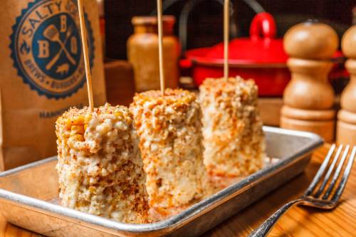 Three corn on the cob pieces coated in cheese and spices, served on a metal tray with wooden utensils in the background.
