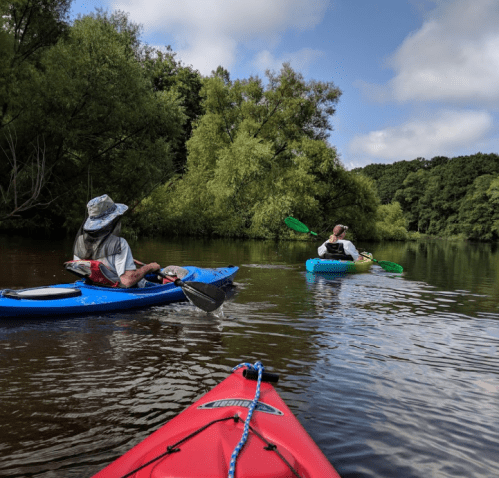 Two people kayaking on a calm river surrounded by lush greenery under a partly cloudy sky.