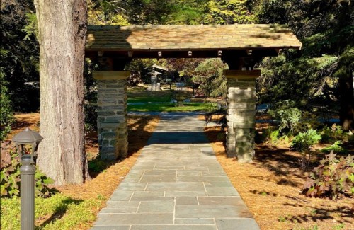 A stone pathway leads through a rustic archway, surrounded by trees and greenery in a serene garden setting.