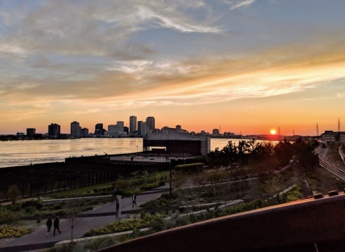 Sunset over a city skyline with a river in the foreground, featuring silhouettes of people and lush greenery.