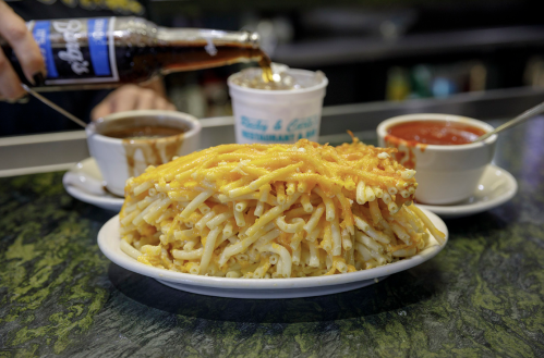 A large plate of cheesy macaroni sits in front of two bowls of sauce, with a bottle of soda being poured into a cup.