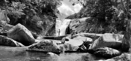 A black and white image of a waterfall surrounded by large rocks and trees, with a person walking near the water.