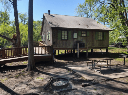 A rustic wooden cabin with green shutters, surrounded by trees, a picnic table, and a fire pit in a sunny outdoor setting.