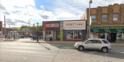 Street view of a pizza shop and a living company storefront, with a car passing by and a bus stop nearby.