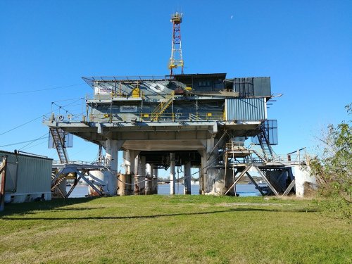 An offshore oil rig stands on stilts over water, surrounded by green grass and a clear blue sky.