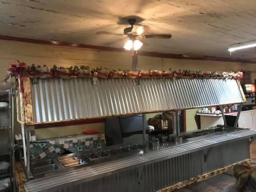A rustic cafeteria serving area with a metal counter, decorated with garlands and a ceiling fan above.