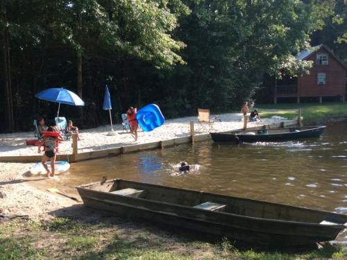 Children playing by a sandy beach near a lake, with boats and umbrellas in a wooded area.