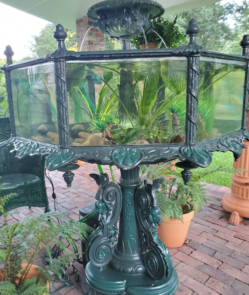 A decorative fountain with a glass tank filled with water and plants, surrounded by greenery and brick flooring.