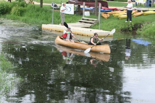 Two people paddle a canoe on a calm waterway, surrounded by greenery and kayaks on the shore.