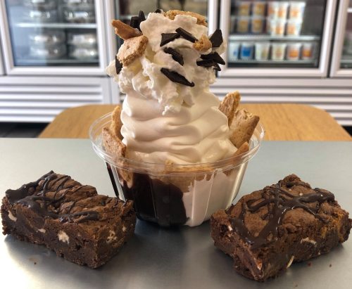 A dessert cup with soft serve ice cream topped with whipped cream, cookie crumbles, and two chocolate brownies on the side.