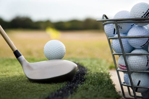 A golf club poised to strike a golf ball near a basket filled with more golf balls on a grassy field.