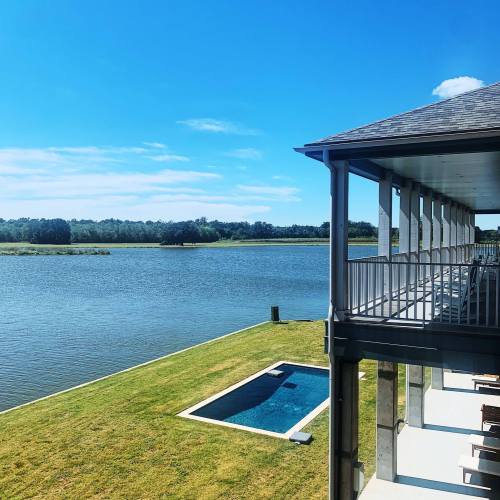 A serene view of a river with a grassy lawn and a small pool beside a modern house under a clear blue sky.