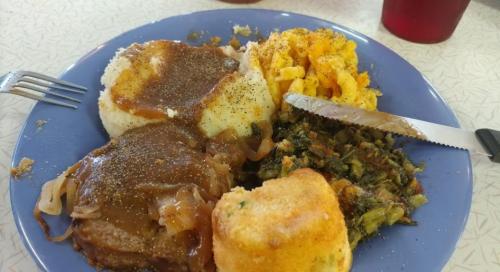 A blue plate with meatloaf, mashed potatoes, macaroni and cheese, greens, and cornbread, garnished with pepper.
