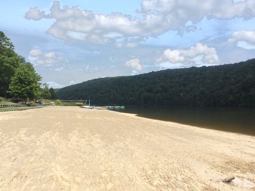A sandy beach by a calm lake, surrounded by green hills and a partly cloudy sky. Volleyball net in the distance.