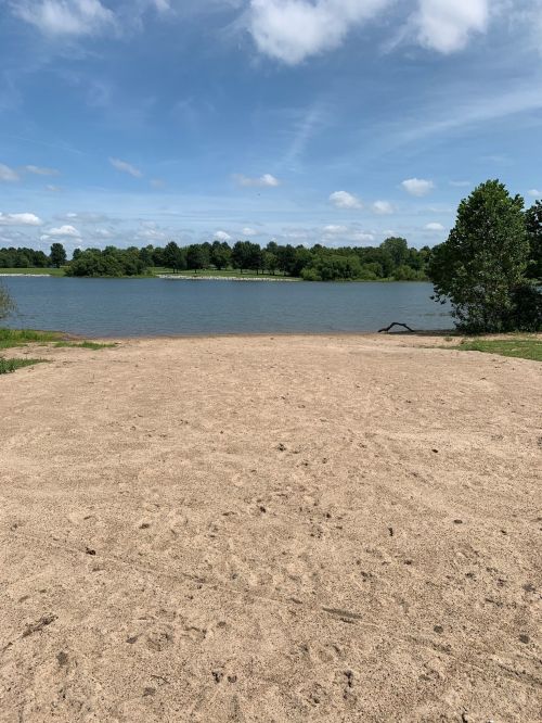 A sandy shore by a calm lake, surrounded by green trees under a blue sky with scattered clouds.