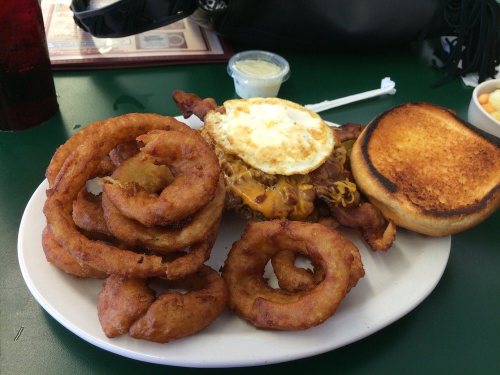 A plate of food featuring a sandwich with bacon, cheese, and a fried egg, accompanied by crispy onion rings and toast.