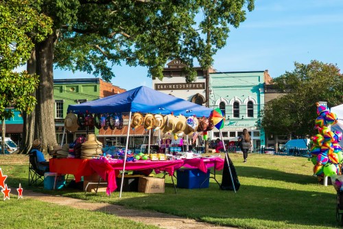 A vibrant market scene with a blue tent displaying hats and colorful items, set in a park with historic buildings in the background.