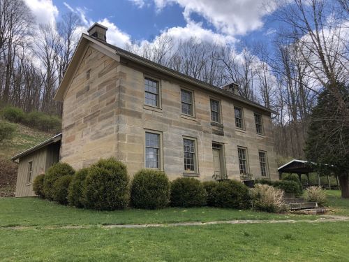 A two-story stone house surrounded by greenery and trees under a partly cloudy sky.