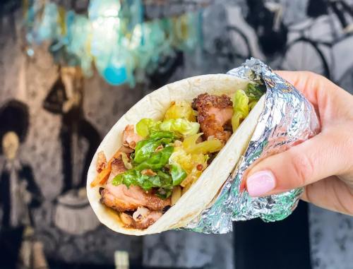 A hand holding a foil-wrapped taco filled with grilled chicken, lettuce, and other toppings against a stylish background.
