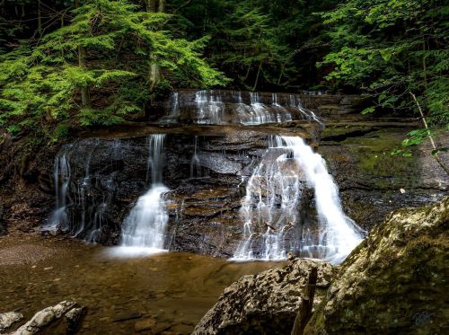 A serene waterfall cascades over rocky ledges, surrounded by lush green trees in a tranquil forest setting.
