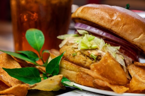 A close-up of a burger with lettuce and onion, served with crispy chips and a drink in the background.