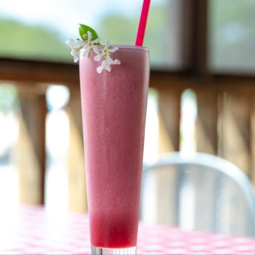 A tall, pink smoothie in a glass with a straw and a flower garnish, set on a table with a blurred background.