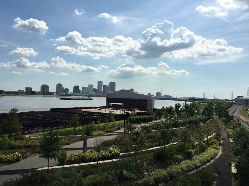 A scenic view of a river with a city skyline in the background, featuring greenery and blue skies with clouds.