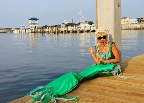 A woman in a green mermaid costume sits on a dock by the water, smiling and pointing playfully.