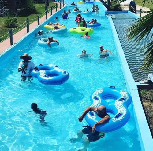 A busy lazy river with people floating on tubes and swimming in a bright blue pool under sunny weather.