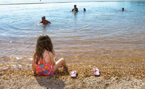 A young girl in a swimsuit sits on the shore, watching people swim in the water on a sunny day.
